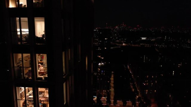 Rising shot of tall residential building at night. Tight flight along lighted windows with views of interiors. London, UK