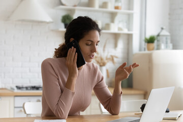 Confident young woman making phone call, sitting at table with laptop in kitchen, businesswoman...