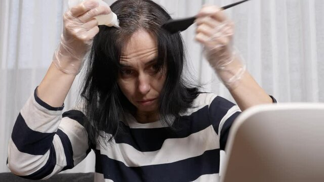 Hair coloring at home. Middle age woman dyeing hair using black brush. Woman colouring her dark hair with regrown gray roots