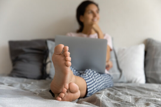 Close up feet of dreamy barefoot woman sitting relaxing on cozy comfortable bed at home with laptop, beautiful young female dreaming, visualizing, enjoying lazy leisure time alone, rest and pedicure