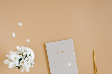 Blogger or freelance workplace. Beige notebook with pen, white flowers out of focus on beige table. Copy space, flat lay