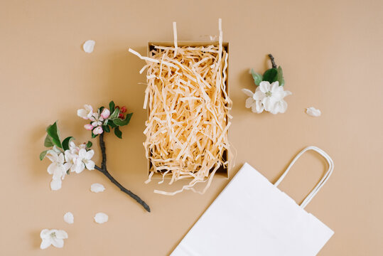 Mockup of an open gift box with peach colored paper shavings, white paper bag and apple flowers on a beige background. Flat lat, top viw