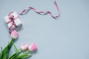 A white gift box with a pink satin bow and a bouquet of pink tulips on a blue background. Copy space