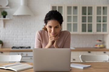 Smiling woman looking at laptop screen, working or studying online in modern kitchen at home, interested positive young female watching webinar or video training, reading good news in message