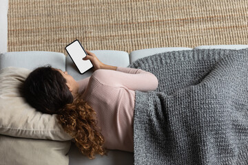 Top view of woman covered with blanket relaxing with smartphone, empty white screen mockup, young...