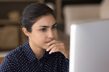 Focused pensive Indian professional working at computer from home, reading email letter, watching webinar. Young business woman thinking over online project, making decision, touching chin