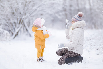 Happy young mother and little daughter playing with snowballs at white snow covered nature park after blizzard. Lovely fun moment. Spending time together in beautiful cold winter day. Side view.