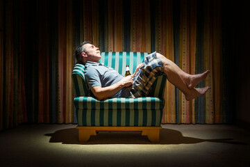 Elderly man with a bottle of beer in a dark room in armchair under light of a lamp
