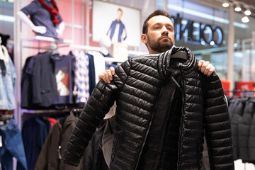 Fototapeta na wymiar Adult handsome brunet unshaven man chooses warm black jacket in a clothing store in a shopping mall. Outerwear and shop