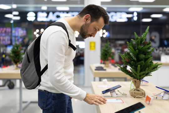 Photo of an adult handsome doubtful thinking brunette man with stubble in a white sweatshirt chooses a mobile phone in an electronic store in a shopping mall. Shop concept
