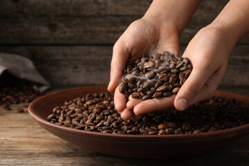 Woman taking pile of aromatic roasted coffee beans from bowl at wooden table, closeup