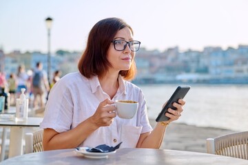 Mature woman in outdoor sidewalk cafe on seafront with cup of coffee using smartphone