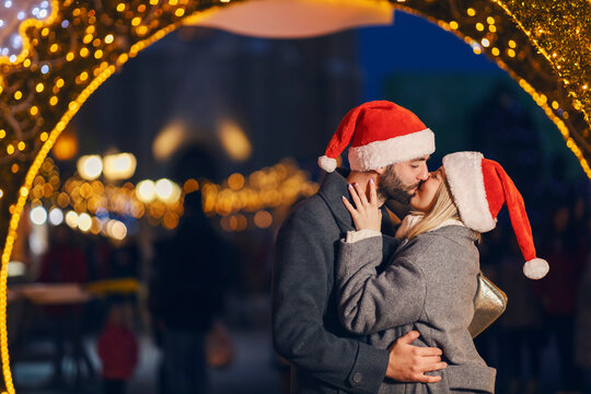 Christmas people kissing outdoors on New Year's eve. A young couple in love with Santa hats on heads standing on the street and kissing on the lips during the Christmas holidays.