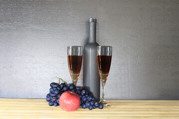 Glass of red wine near wine bottle next to apple and bunch of grapes on gray background