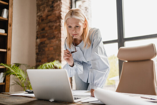Concentrated caucasian middle-aged mature businesswoman ceo boss typing on laptop, working at office desk with documents, searching surfing web online