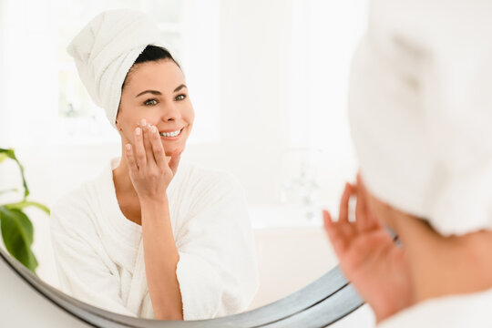 Anti-age anti-wrinkle effect of moisturizing creme. Caucasian mature middle-aged woman in turban spa bathrobe applying beauty product on face skin.