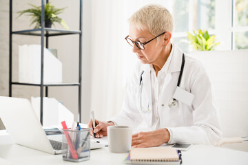 Caucasian senior elderly mature female woman doctor nurse in white coat writing prescriptions for medicines drugs for curing ill patients