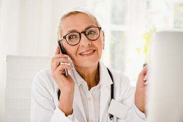 Smiling mature senior caucasian female family doctor in white coat talking on smart phone with patients, telemedicine on distance, pandemic concept.