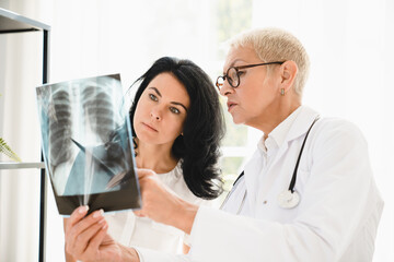 Caucasian senior female doctor general practitioner showing x-ray lungs photo to female patient,...