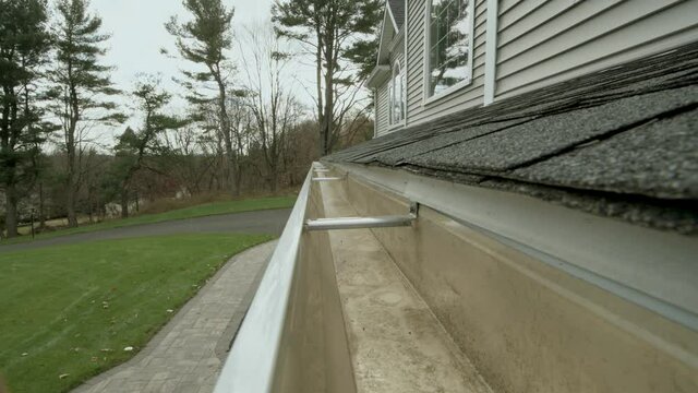 Zoom out Cleaned K style white aluminum gutter that has been cleaned a view looking down the length of the gutter trough and most of the leaves have fallen on an overcast day Gray shingles vinyl 