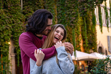 Young couple traveling to Rome. Beautiful man with long hair hugs his girlfriend who is laughing. In the background a street in the historic center with a building covered with ivy.