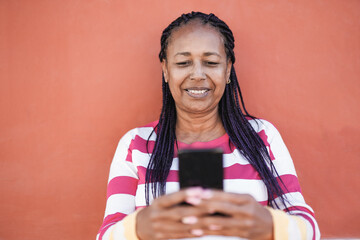 Trendy elderly african woman using mobile phone in the city with red background