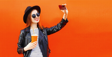 Portrait of stylish smiling young woman taking a selfie picture by smartphone with cup of juice on orange background