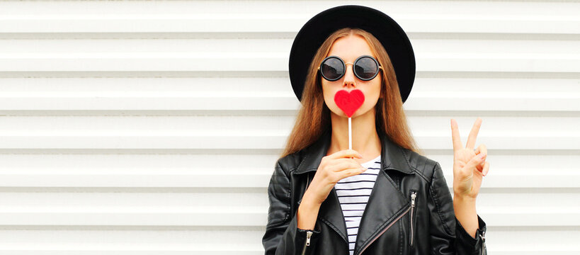 Fashionable portrait of stylish young woman with red heart shaped lollipop blowing her lips sending sweet air kiss wearing a black round hat, leather jacket on white background