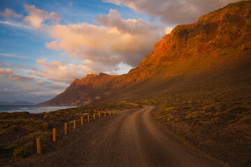 Sunset near Famara Beach, Lanzarote, Spain. A trail along the ocean and the cliffs. Some clouds in a blue sky.