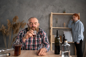 alcoholism in the family, a sad drunk husband pours alcohol into a glass and a sad relaxed wife in the background, alcohol abuse at home. Addiction and depression. C