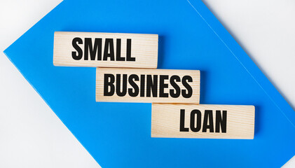 There is a blue notebook on a light gray background. Above are three wooden blocks with the words SMALL BUSINESS LOAN