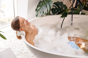 Relaxed woman looking happy while taking bubble bath in her beautiful bathroom decorated with plants. Spa resort, wellness concept