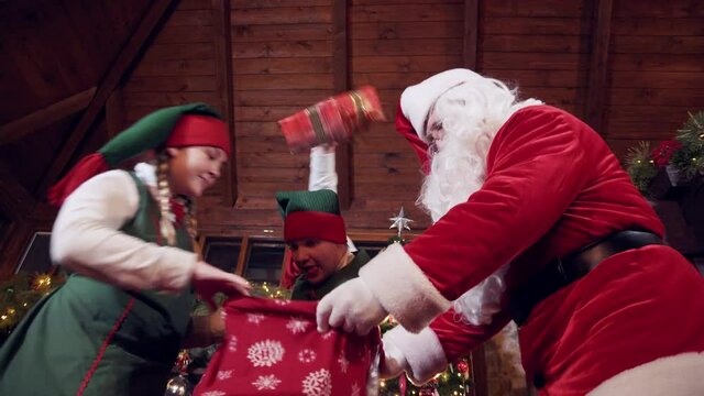 Santa Claus and elves pack red sack with gifts. Cheerful elves and Santa pack red bag with Christmas presents for kids. View from below.