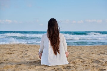 Fototapeta na wymiar Brunette in white with long hair sitting on sandy beach with her back looking at sea