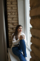 A beautiful girl in a white sweater is sitting on the windowsill and drinking coffee.