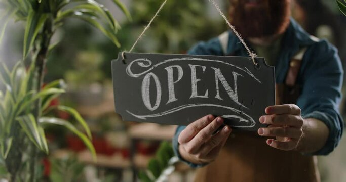 Flower shop employee flipping sign on glass entrance door from closed to opened, starting workday of greenhouse. Small business people 4k footage