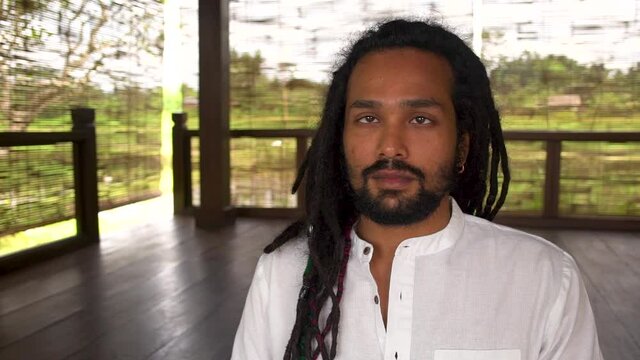 Close-up of indian man with dreadlocks do meditation practice, young man wearing white shirt sitting at yoga place, he closes his eyes and plunges into meditative state. Dollyshot.