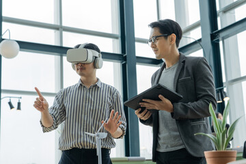Two young happy Asian businessmen are relaxing during break by one man wearing a virtual reality simulator headset, another one holding a tablet, playing game together in office creative room space.