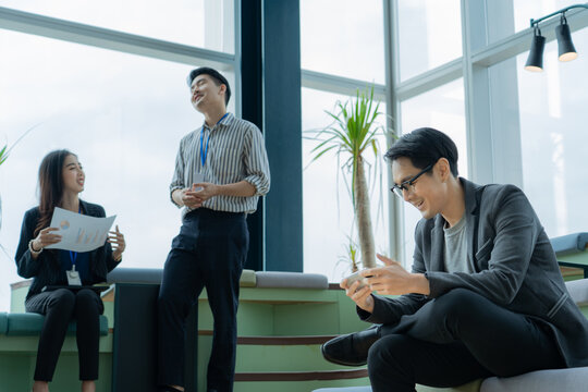 Group of young happy Asian business people, two men and one woman chilling and relaxing during break by talking together and enjoy playing game on mobile in a creative room space at a modern office.