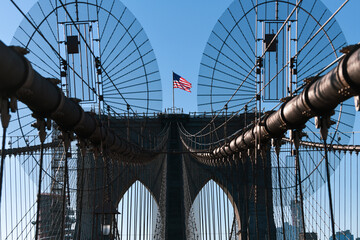 Obraz premium The national flag of United States of America winding on top of Brooklyn Bridge landmark from Manhattan. One of the most visited places in America. Symbol of patriotism.