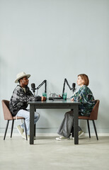 Vertical full length portrait of two people speaking to microphones while recording podcast in...