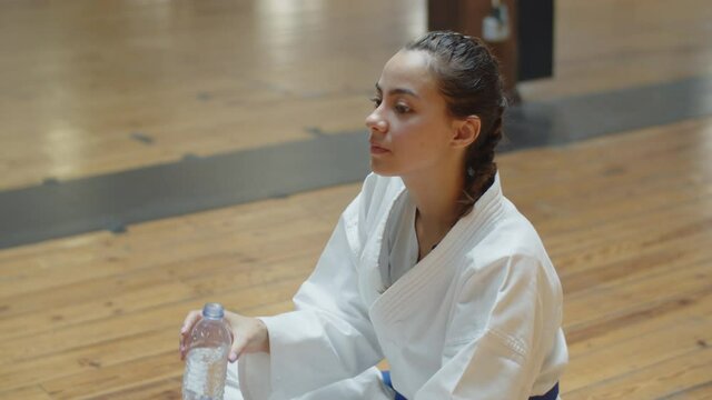 Side view of karateka sitting on floor and drinking water. Medium shot of pensive girl in kimono having break, resting after karate workout, recovering strength. Martial arts, relaxation concept