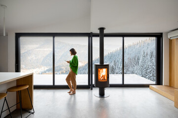Woman standing with phone near fireplace at modern living room with great view on snowy mountains....