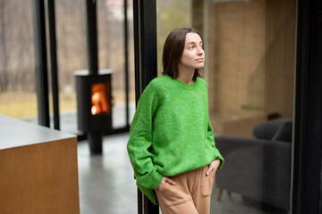 Young woman stands at window of a modern house on nature and look away. Concept of comfort and...