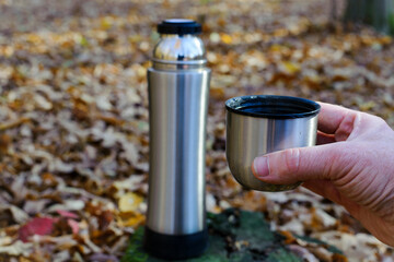 cup with tea in hand. a thermos of tea, hot drink. thermos on an old tree stump. The concept of tourism, survival and hunting in the autumn in forest. dry leaves in the park. rest at nature.