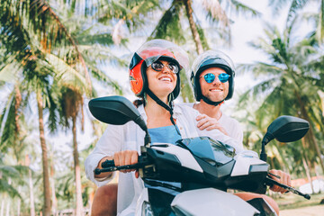 Happy smiling couple travelers riding motorbike scooter in safety helmets during tropical vacation...