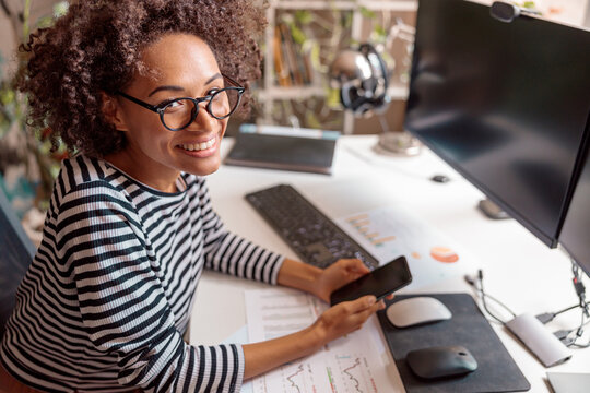 Joyful multiethnic lady holding smartphone and smiling while sitting at the table with papers and desktop computer