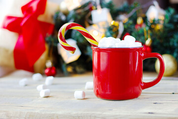 cozy winter hot chocolate drink with marshmallows and lollipop in red mug on background of christmas decor, cute bright card with hot milk in a mug on a wooden background