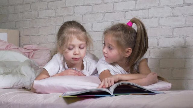 Children read a book, look at pictures, study while lying in bed. Girls in the bedroom. Two preschooler children. Beautiful, funny, cute, smart kids