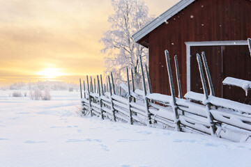 An old red barn and traditional fence in a winter landscape at sunset. A typical Finnish rural landscape in winter.